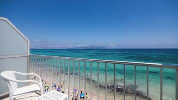 Un balcon avec des fauteuils en osier blanc et une table donnant sur une plage où l’eau est d’un bleu turquoise éclatant et où l’on peut voir des gens se baigner.