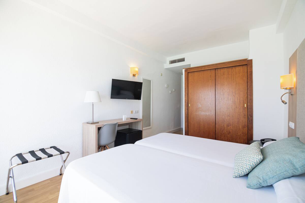 Quarto de hotel com cama, escrivaninha com cadeira, televisão e guarda-roupa.