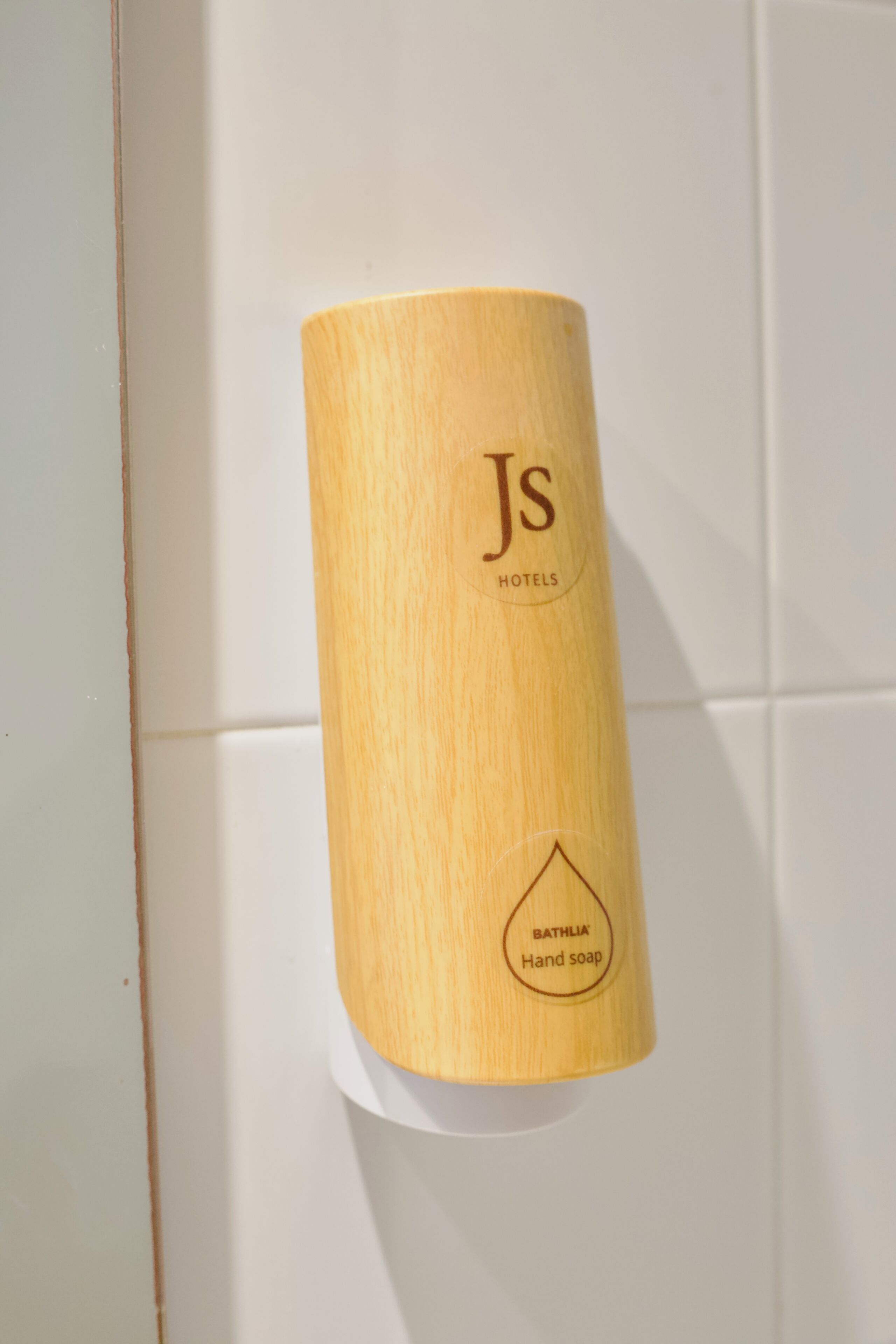 Een cilindrische zeepdispenser met het logo 'Js Hotels' en de tekst 'Bathlia Hand Soap' op een witte betegelde muur.