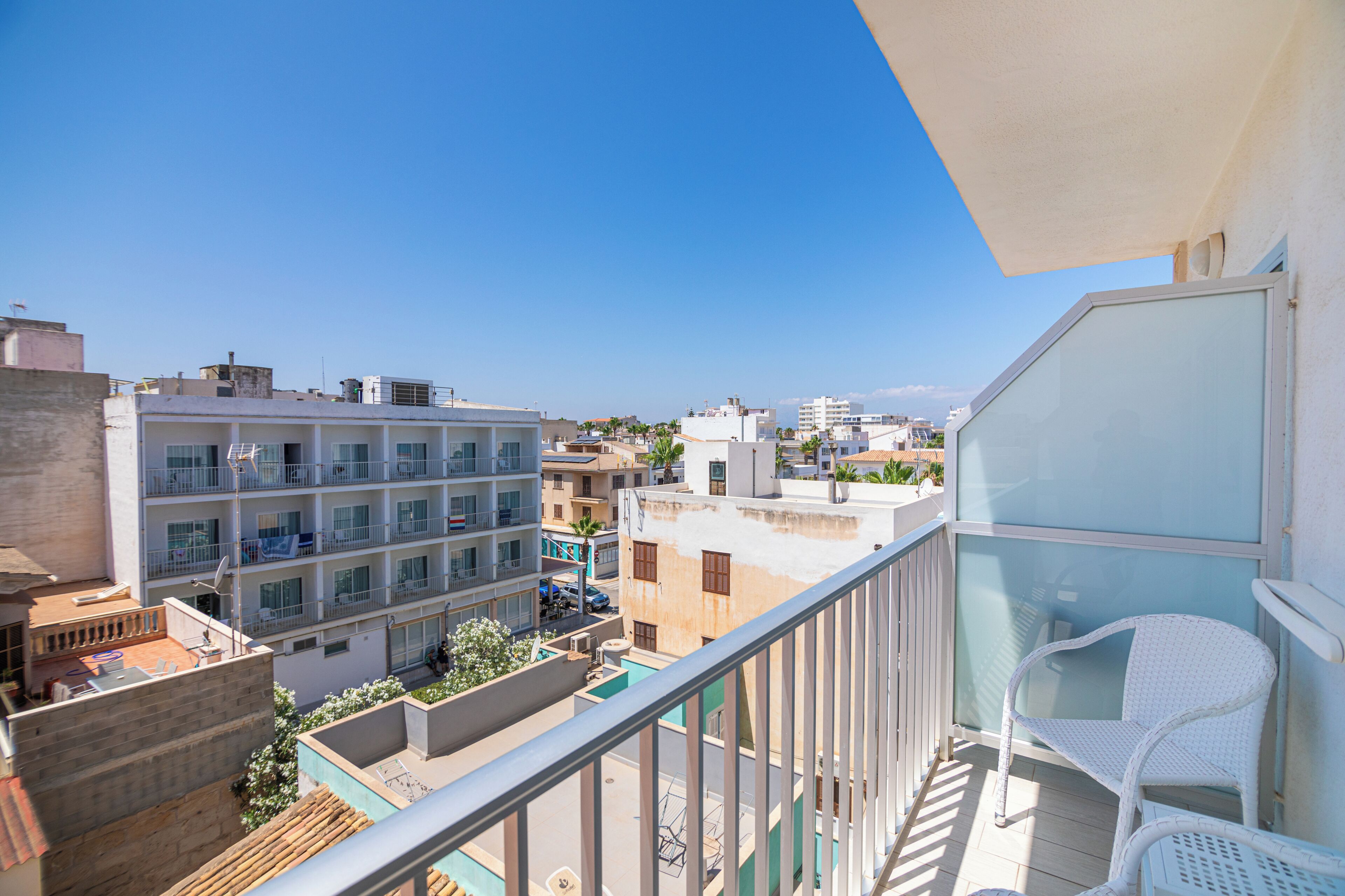 A balcony with a glass railing overlooking a cityscape, featuring a building with multiple balconies and a clear blue sky.