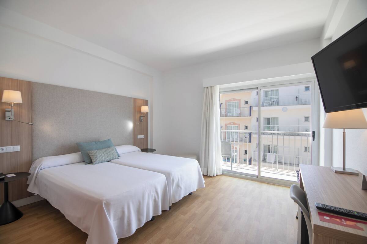 Quarto de hotel com duas camas, uma escrivaninha, televisão e janela com vista para edifícios.