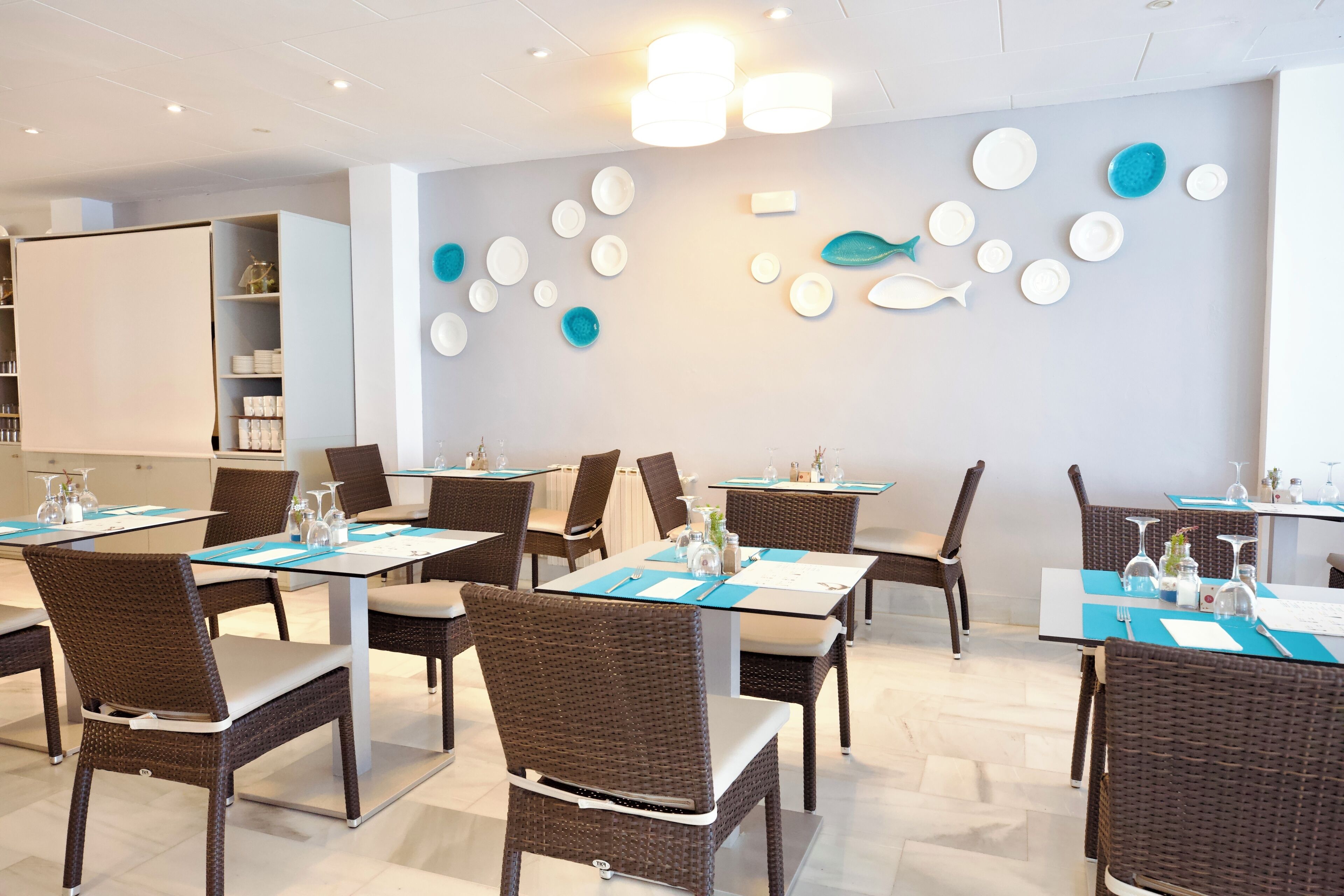 Un comedor con mesas dispuestas para cuatro personas, sillas de mimbre y una pared decorada con platos.