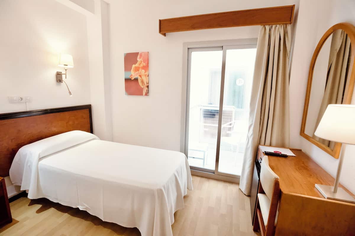 Quarto de hotel com cama, escrivaninha, luminária, pintura e janela com cortinas.