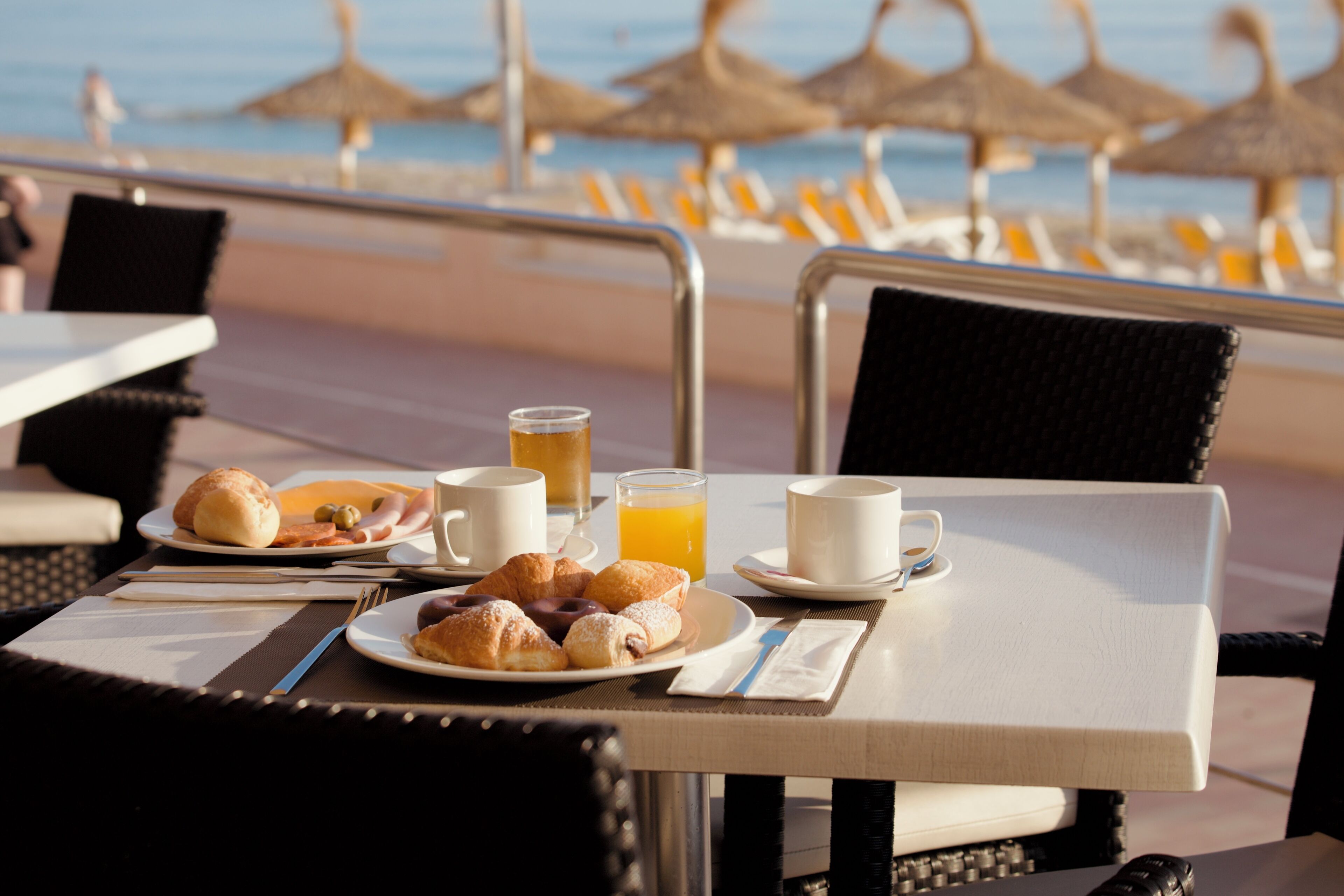 Una mesa con alimentos para el desayuno, un vaso de jugo de naranja y una taza, con vista a una playa con sombrillas.