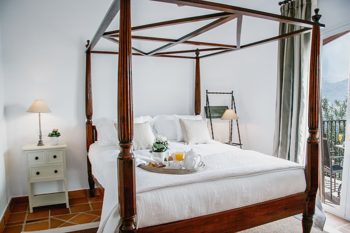 A four-poster bed with a tray of breakfast items, a nightstand with a lamp, and a view of the outdoors.