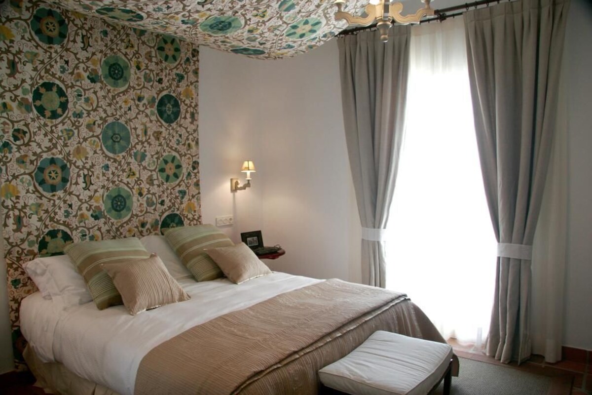 Un dormitorio con papel tapiz estampado, una cama con almohadas, una mesita de noche con lámpara y una ventana con cortinas.