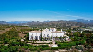 Un grand bâtiment blanc, doté de nombreux balcons, entouré de jardins et de vignobles bien entretenus, et se dressant sur un fond de montagnes.