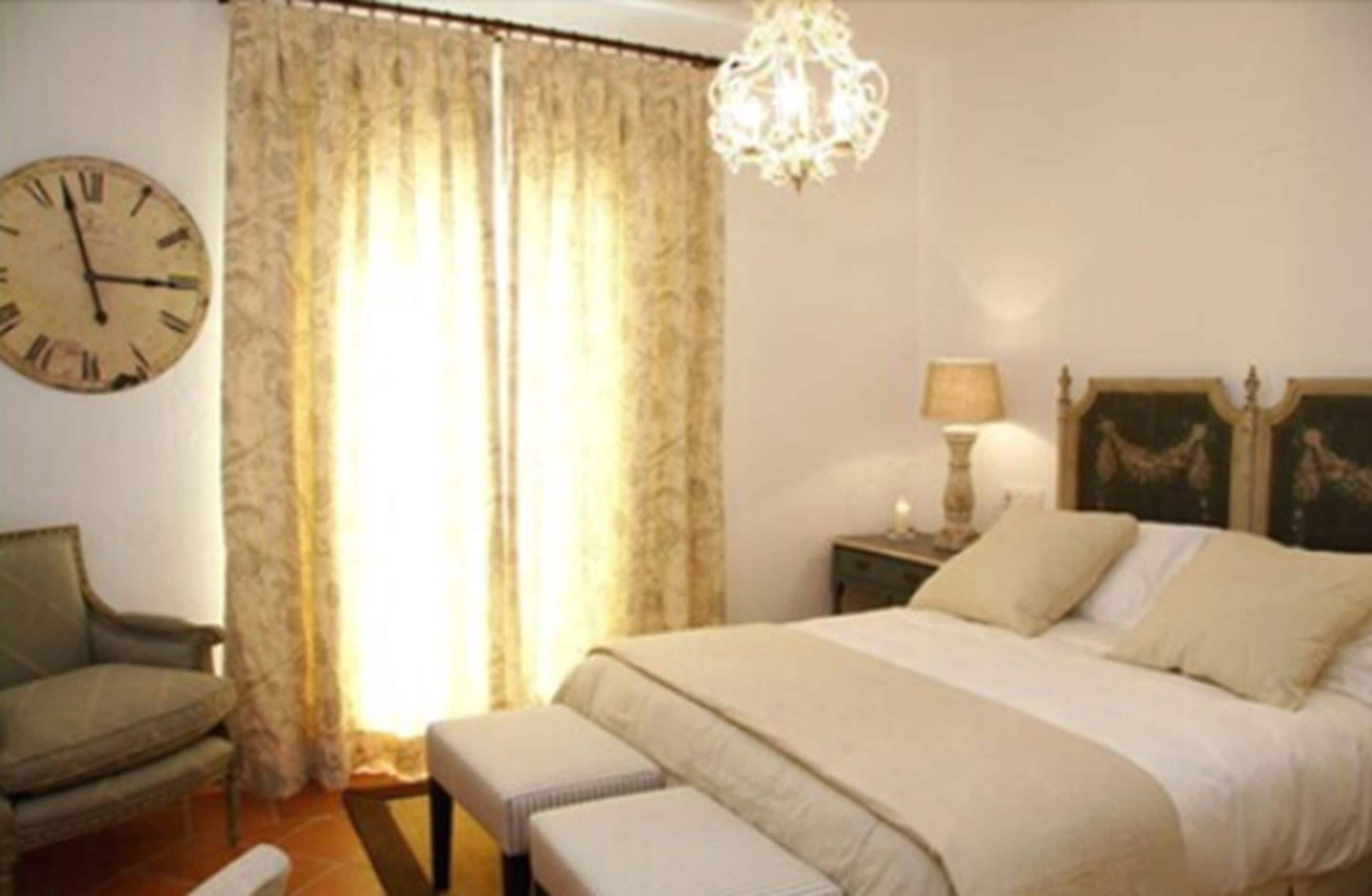 A bedroom with a bed, a chair, a nightstand with a lamp, a clock, and a chandelier.