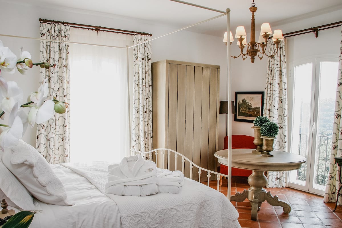 A bedroom with a bed, white curtains, a wooden wardrobe, a chandelier, and a table with potted plants.