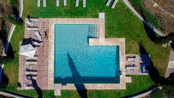 An aerial view of a rectangular swimming pool with a tiled deck surrounding it, equipped with lounge chairs and umbrellas.