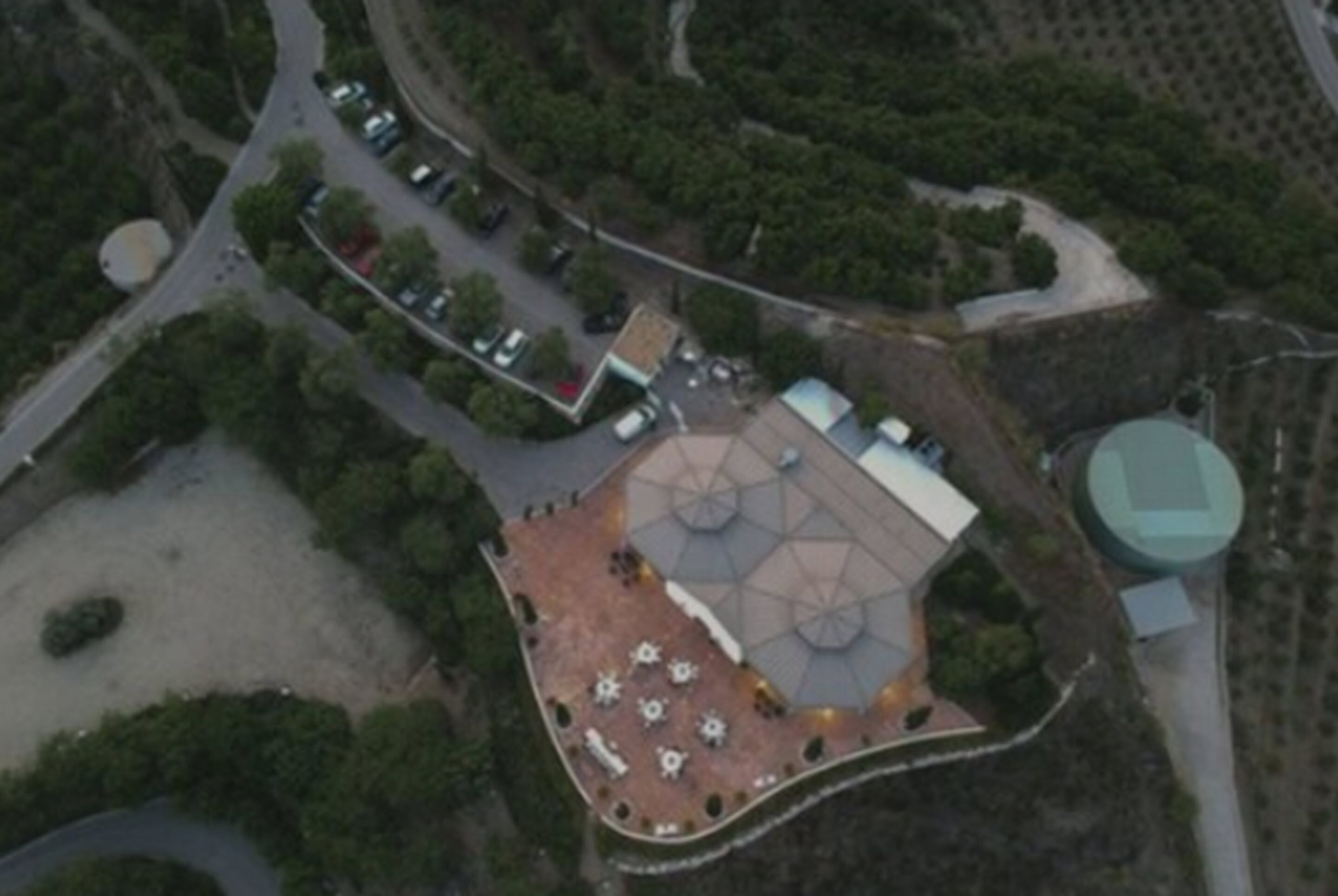 An aerial view of a building with a unique geometric roof structure, surrounded by greenery and a parking area.