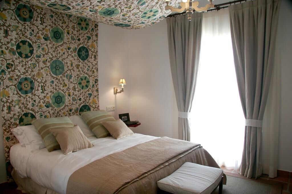 Een slaapkamer met behang met een patroon, een bed met kussens, een nachtkastje met een lamp en een raam met gordijnen.