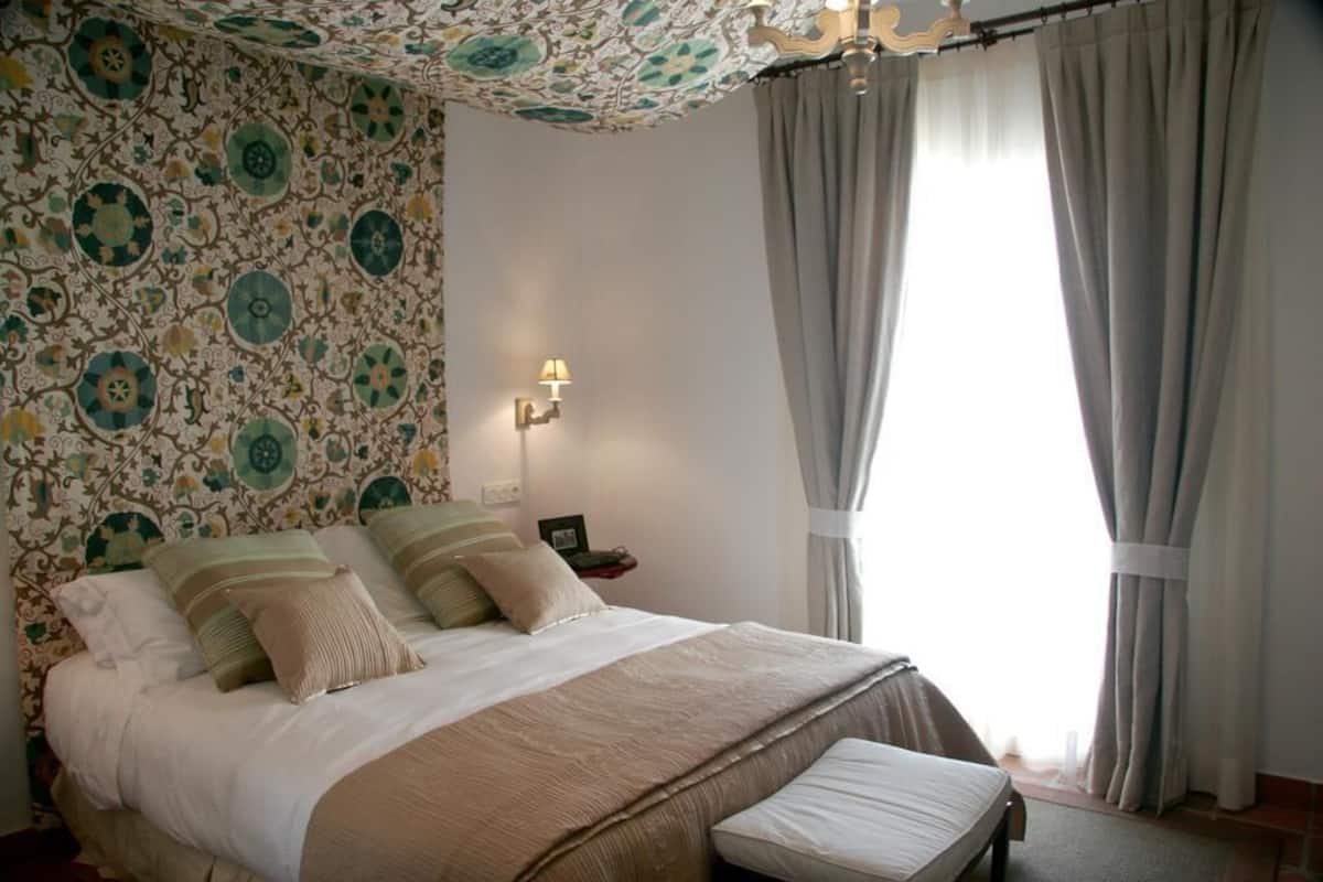 A bedroom with a patterned wallpaper, a bed with pillows, a bedside table with a lamp, and a window with curtains.