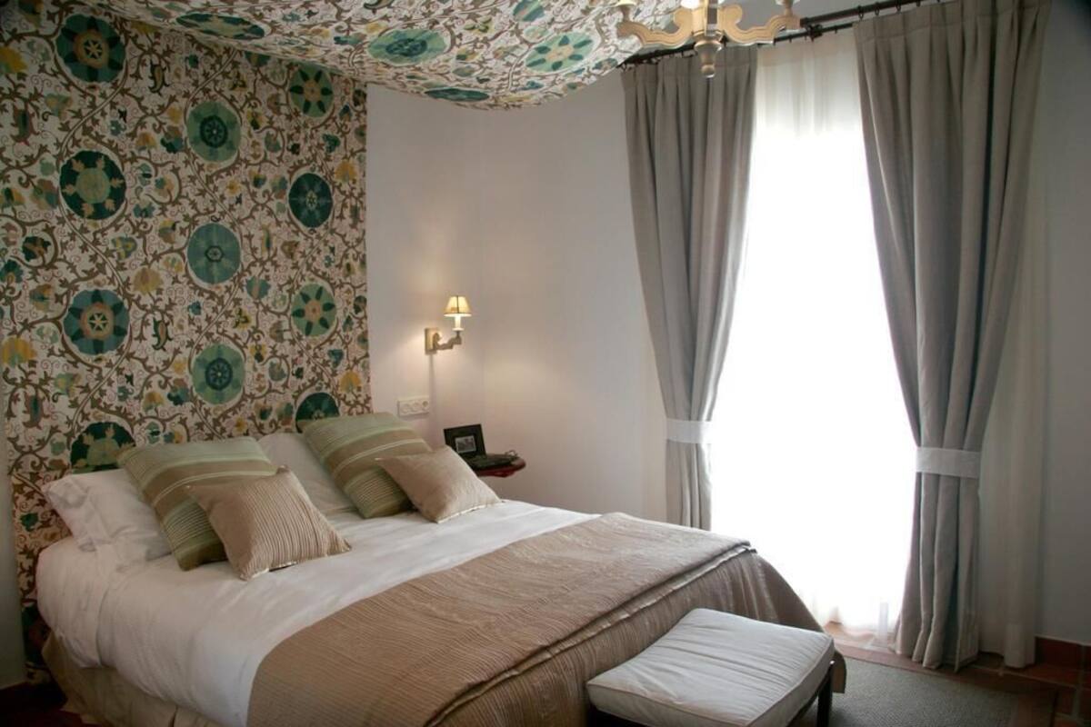 A bedroom with a patterned wallpaper, a bed with pillows, a bedside table with a lamp, and a window with curtains.