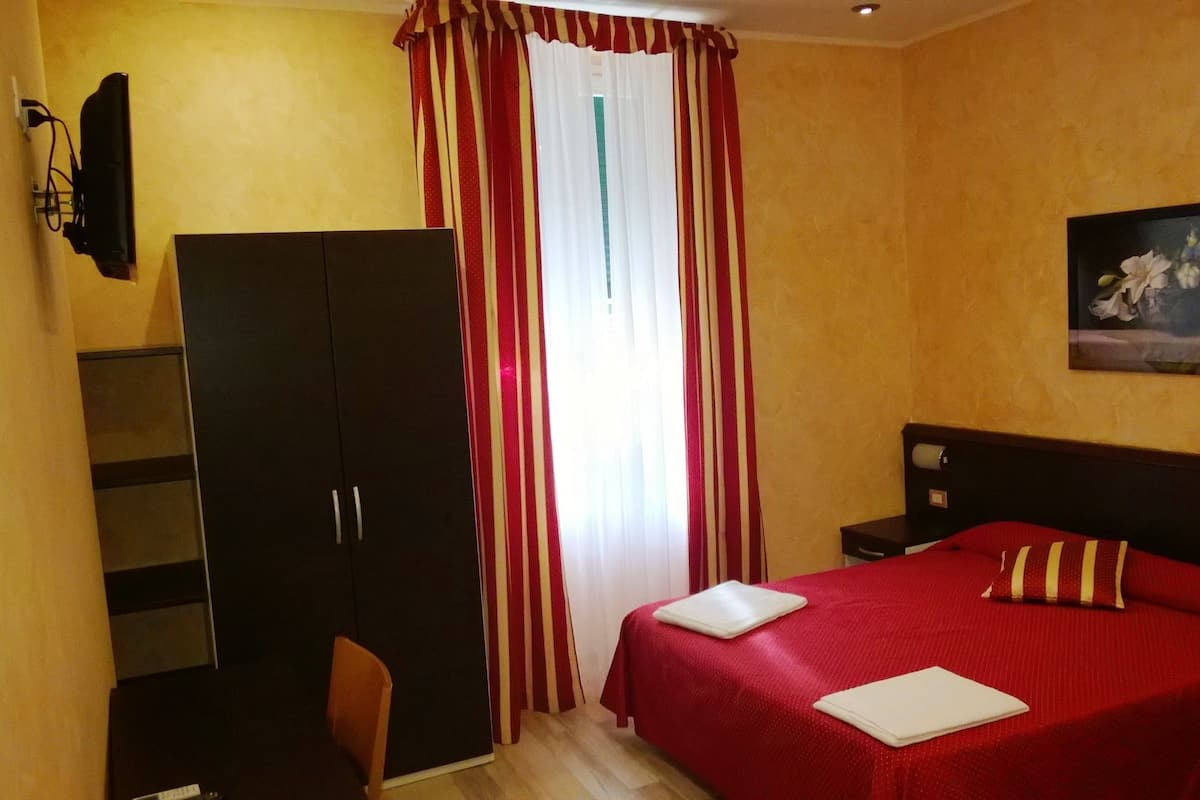 A hotel room with a bed, a desk, a wardrobe, and a painting on the wall.
