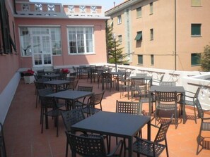 An outdoor patio area with multiple tables and chairs, a pink building with white-framed windows, and a beige building in the background. - Hotel Villa Adele (Celle Ligure)
