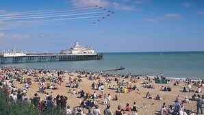 Ein Strand mit einem Pier im Hintergrund und einer Formation fliegender Flugzeuge.
