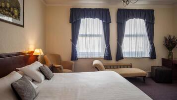 A hotel room with a large bed, two chairs, a bench, a lamp, and two windows with curtains.