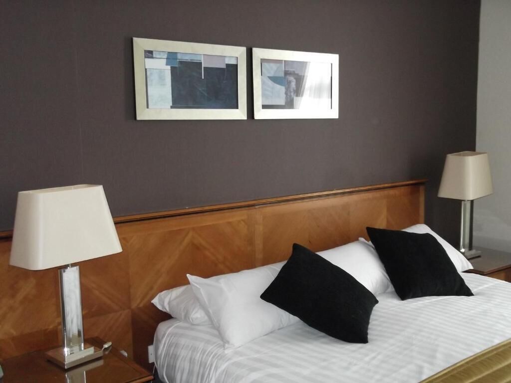 A hotel room with a wooden headboard, a bed with white and black pillows, two bedside lamps, and two framed pictures on the wall.