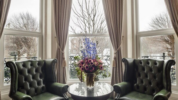Ein Zimmer mit zwei schwarzen Ledersesseln, einem Glastisch und einem Blumenstrauß neben einem großen Fenster mit Vorhängen.
