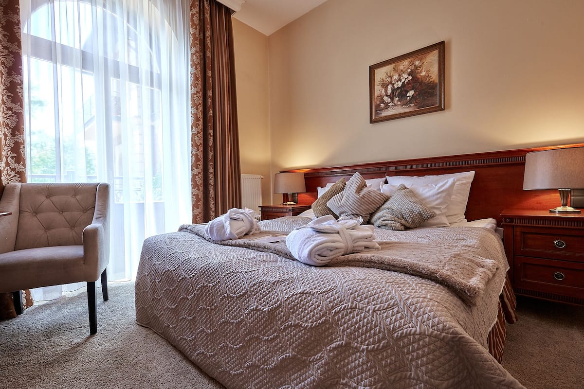 Ein Hotelzimmer mit einem großen Bett, einem Sessel, einem Bild an der Wand und einem Fenster mit Vorhängen.