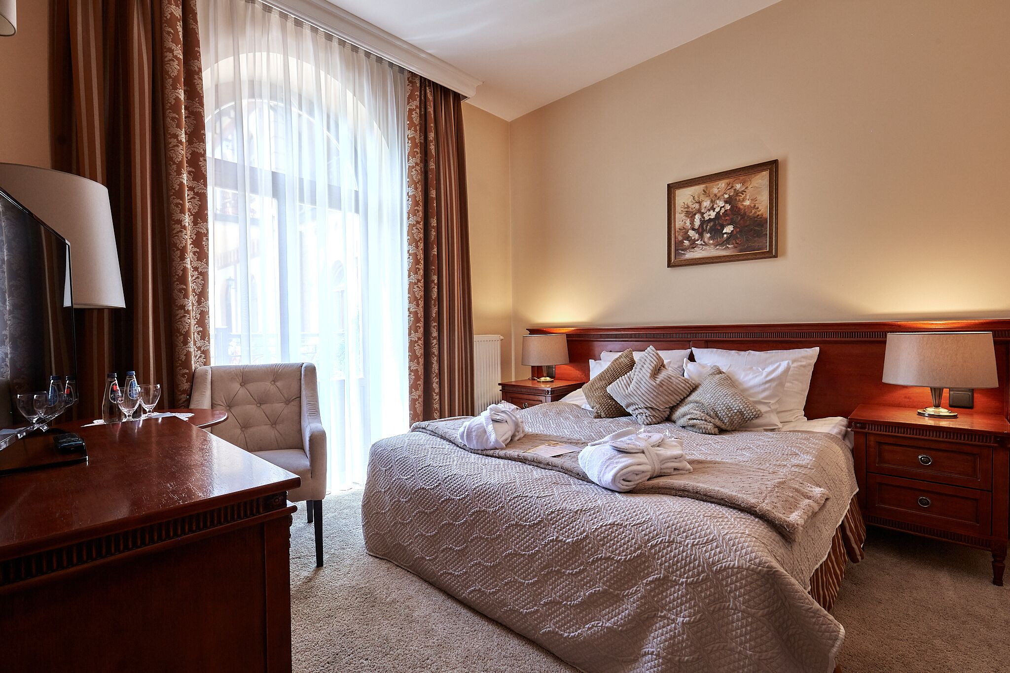 A hotel room with a large bed, a desk with a lamp, a chair, and a painting on the wall.