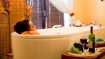 A person relaxing in a bathtub with a bottle of wine and grapes on a table beside the tub.