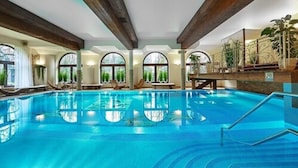 Una piscina interior con agua azul cristalina, ventanas amplias y sillones de descanso.