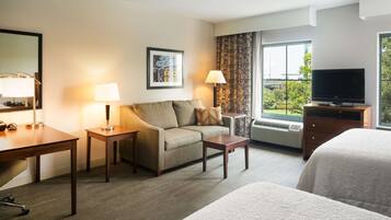 A hotel room with a sofa, a desk, a television, and a large window with a view of greenery.