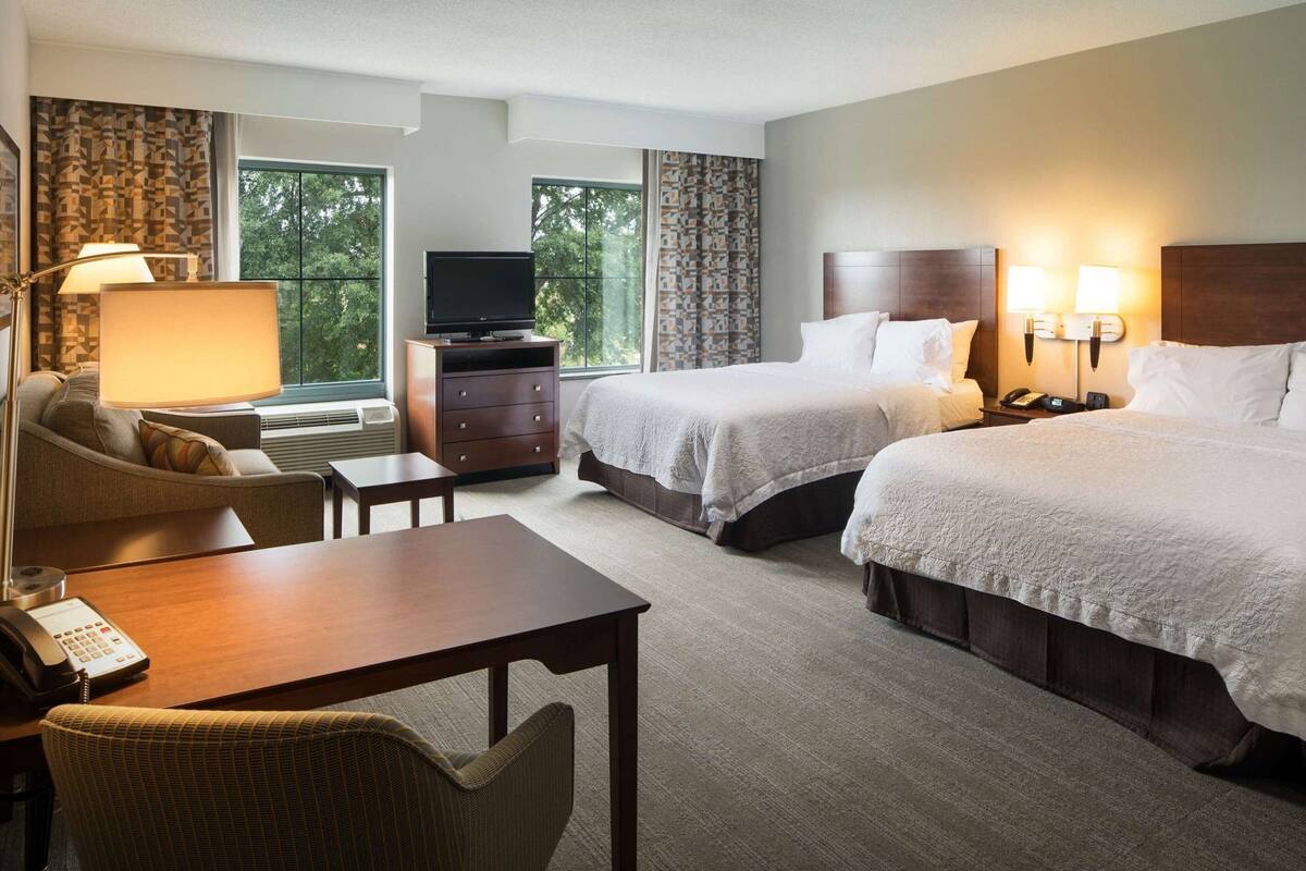 A hotel room with two beds, a desk, a chair, a TV, and a lamp.
