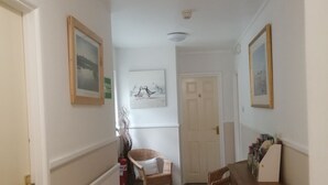 A hallway with a radiator, a wicker chair, a wooden cabinet, and framed pictures on the walls. - Harbourlight Guest House (Saundersfoot)