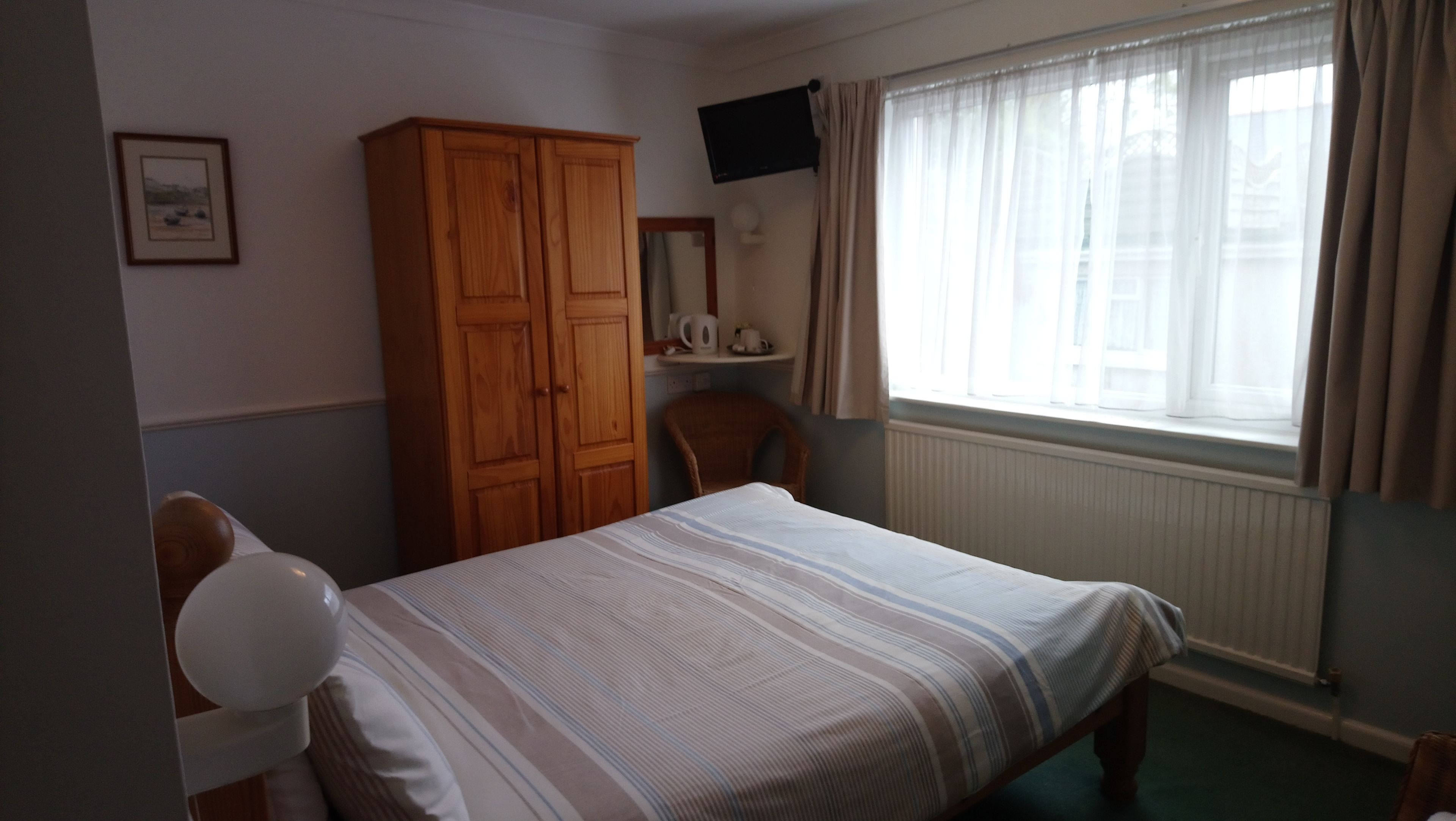 Ein Schlafzimmer mit Bett, Holzschrank, Stuhl, Fernseher und einem Fenster mit Vorhängen.