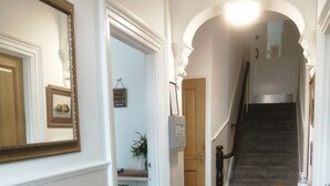 A hallway with a wooden staircase, a fire extinguisher, and a door leading to a room with a shelf and a framed picture. - Harbourlight Guest House (Saundersfoot)