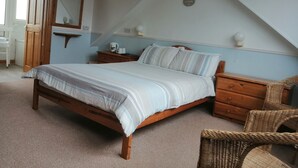 A bedroom with a wooden bed, a nightstand, a wicker chair, and a small table with a lamp. - Harbourlight Guest House (Saundersfoot)