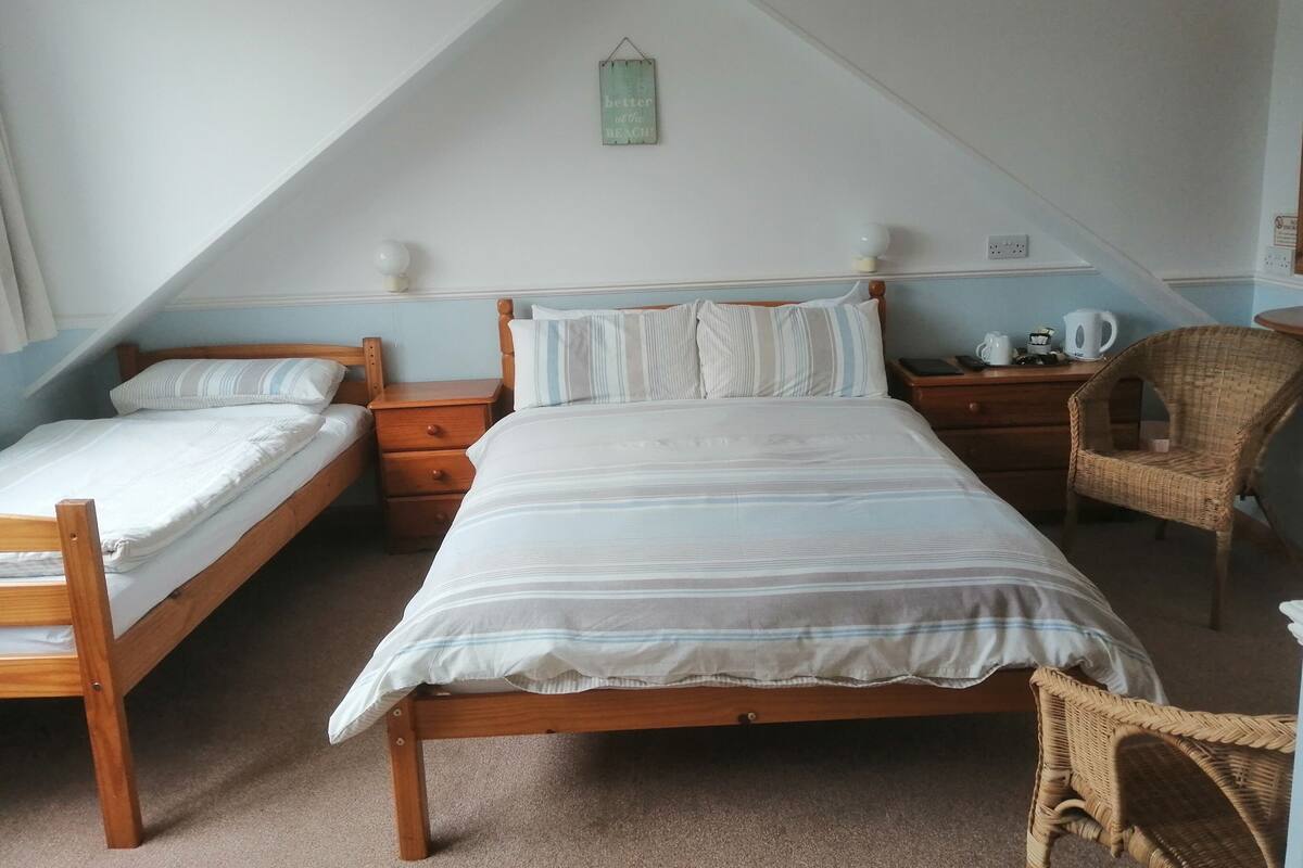 A bedroom with two single beds, a wooden nightstand, a wicker chair, and a small table with a kettle and cups.