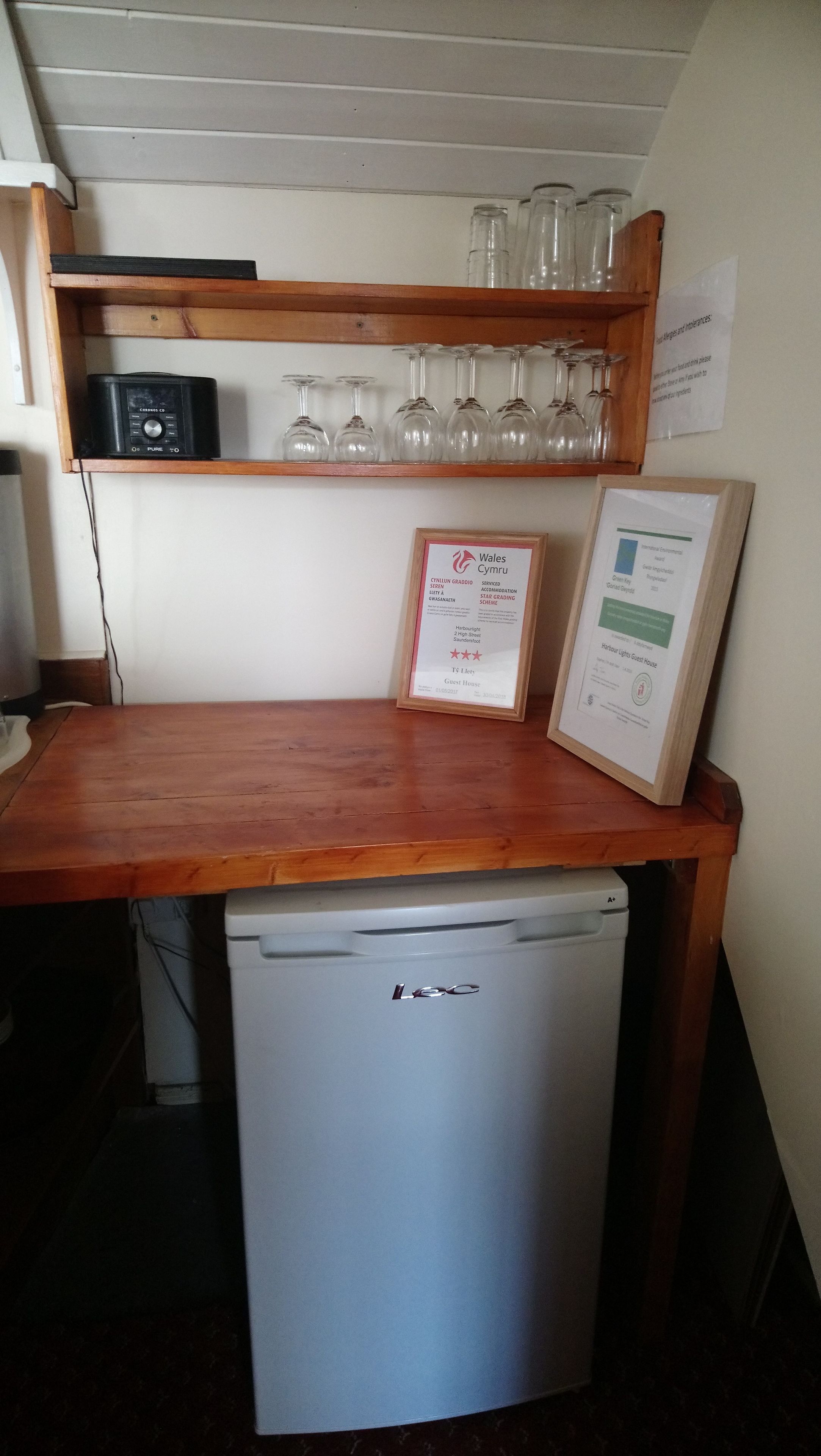 Uma cozinha com bancada de madeira, uma pequena geladeira, uma prateleira com copos de vinho e dois certificados emoldurados na parede.