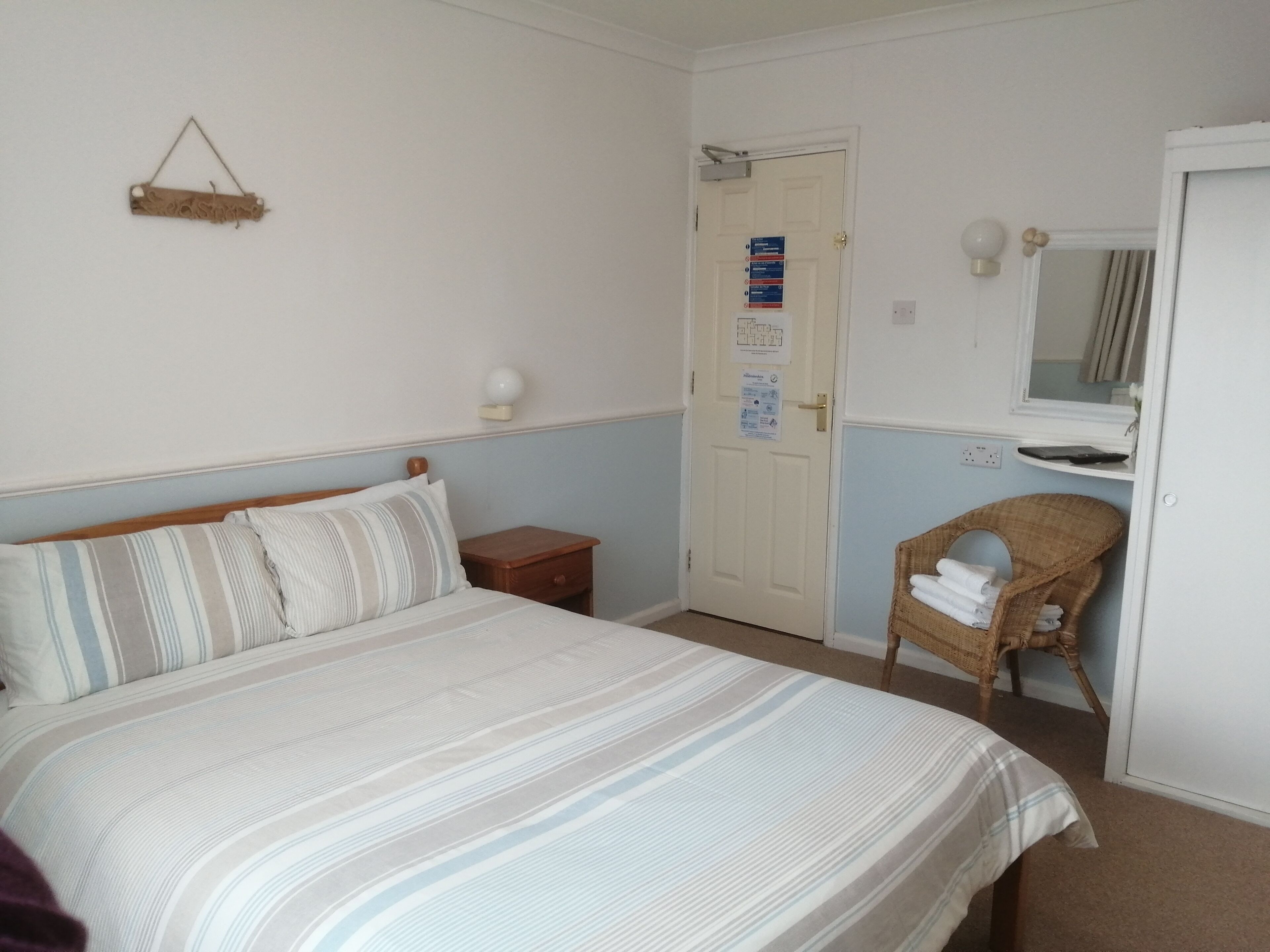 A bedroom with a bed, a nightstand, a wicker chair, a door with a sign, and a mirror.