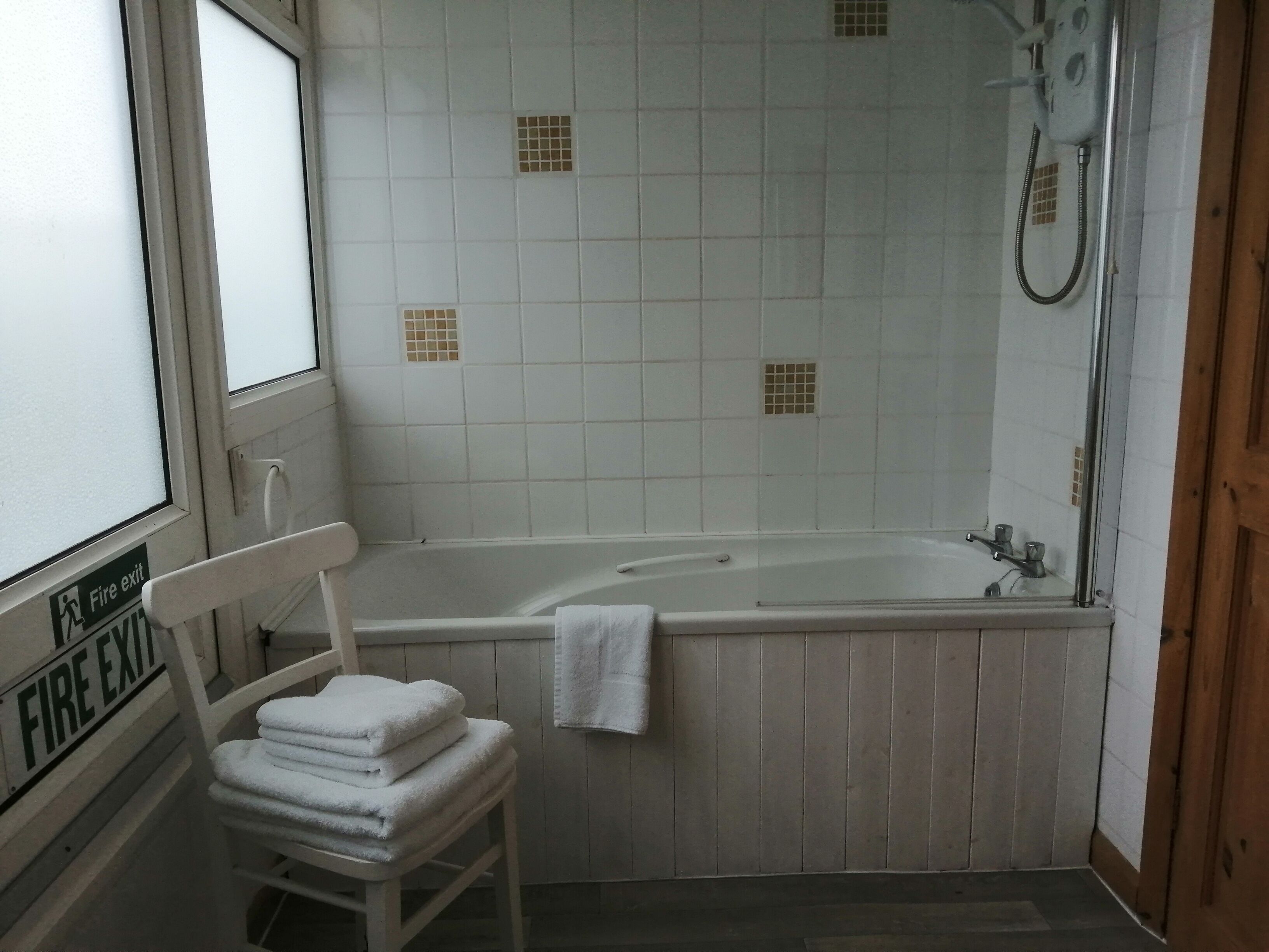 A bathroom with a white bathtub, a shower, and a white chair with towels. There is a fire exit sign on the wall.