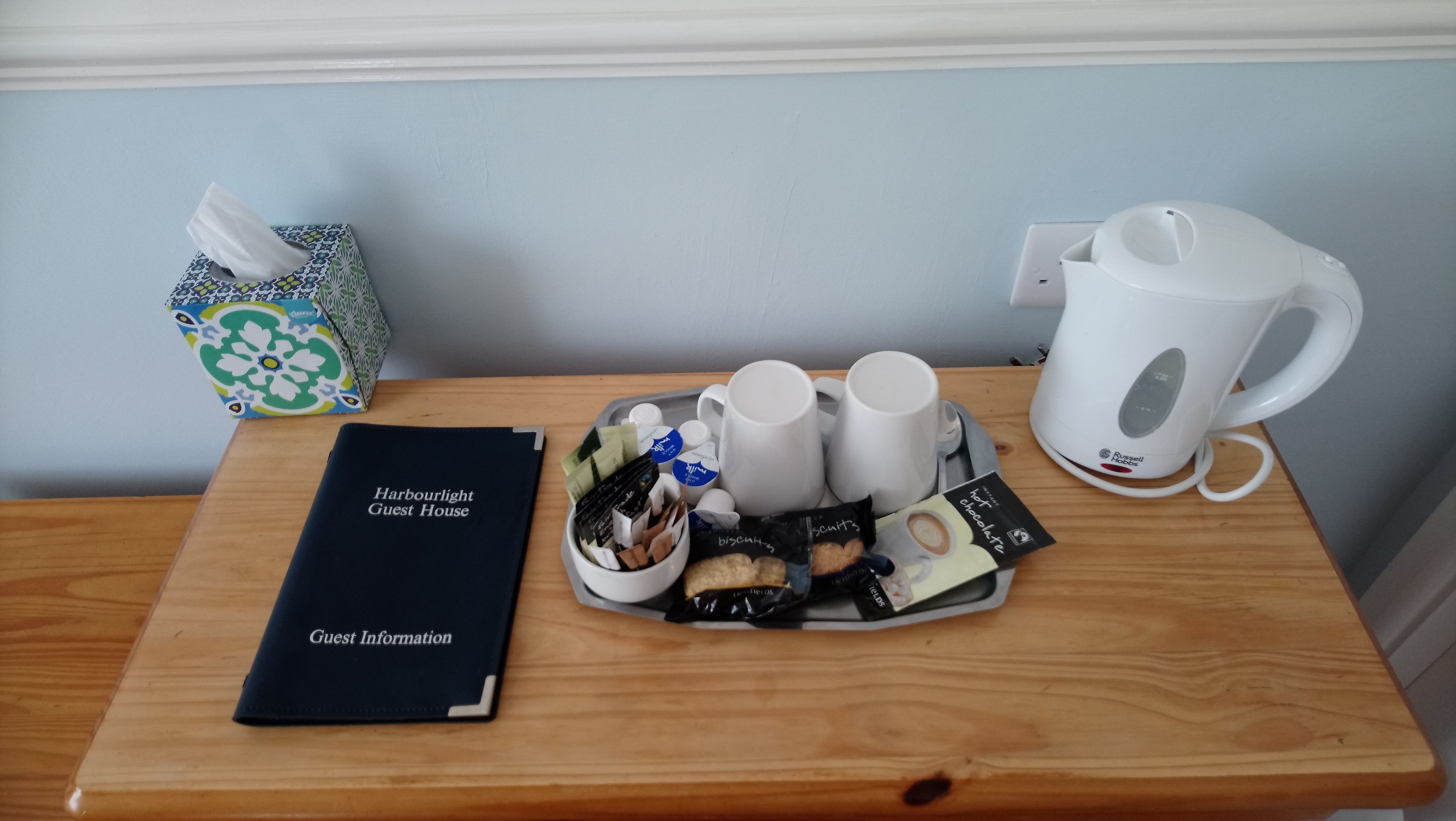A wooden table with a guest information booklet, a tissue box, a tray with biscuits, a mug, and a coffee maker.