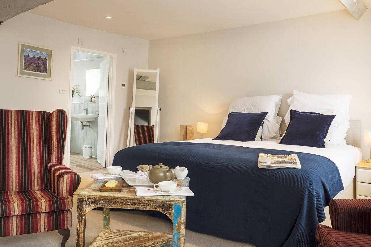 A hotel room with a large bed, a striped armchair, a small table with tea set, and a painting on the wall.