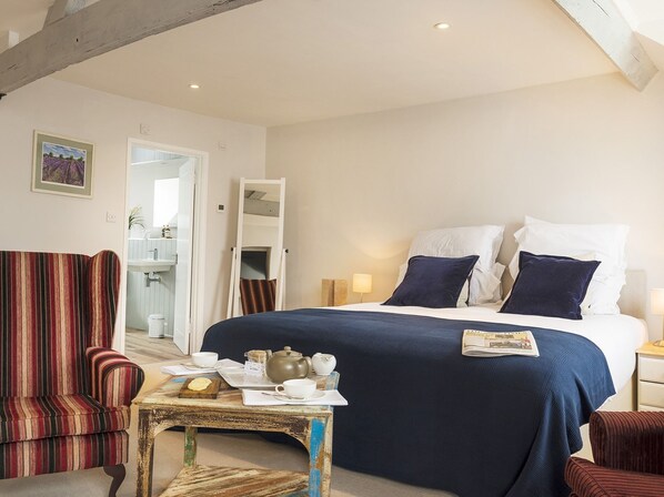 A hotel room with a large bed, a striped armchair, a small table with tea set, and a painting on the wall. - Wild Garlic Rooms (Stroud)