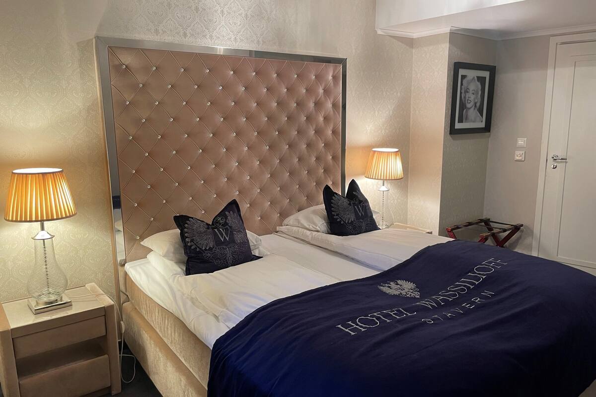 A hotel room with a tufted headboard, two bedside lamps, a bed with a dark blue bedspread, and a framed picture on the wall.