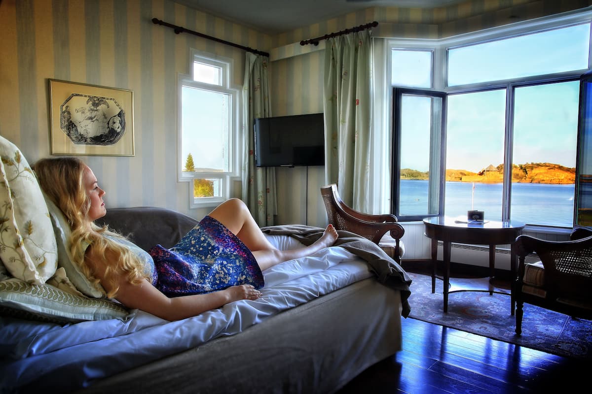 A room with a bed, a TV, a window with a view of the sea, and a table with a chair.