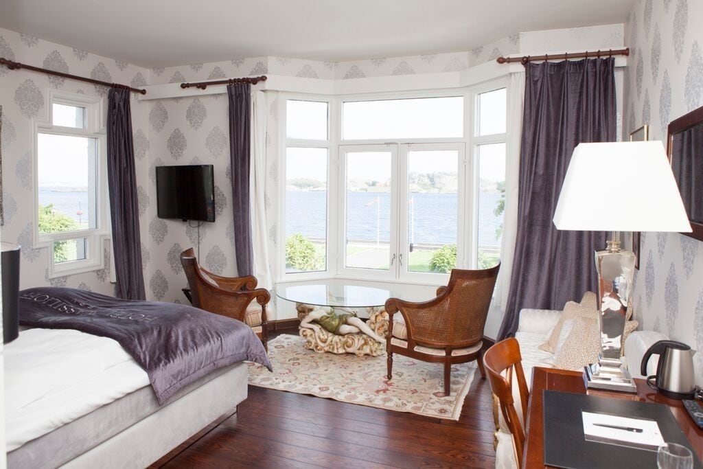 Quarto de hotel com cama, mesa redonda com cadeiras, televisão e vista para o mar através de grandes janelas.