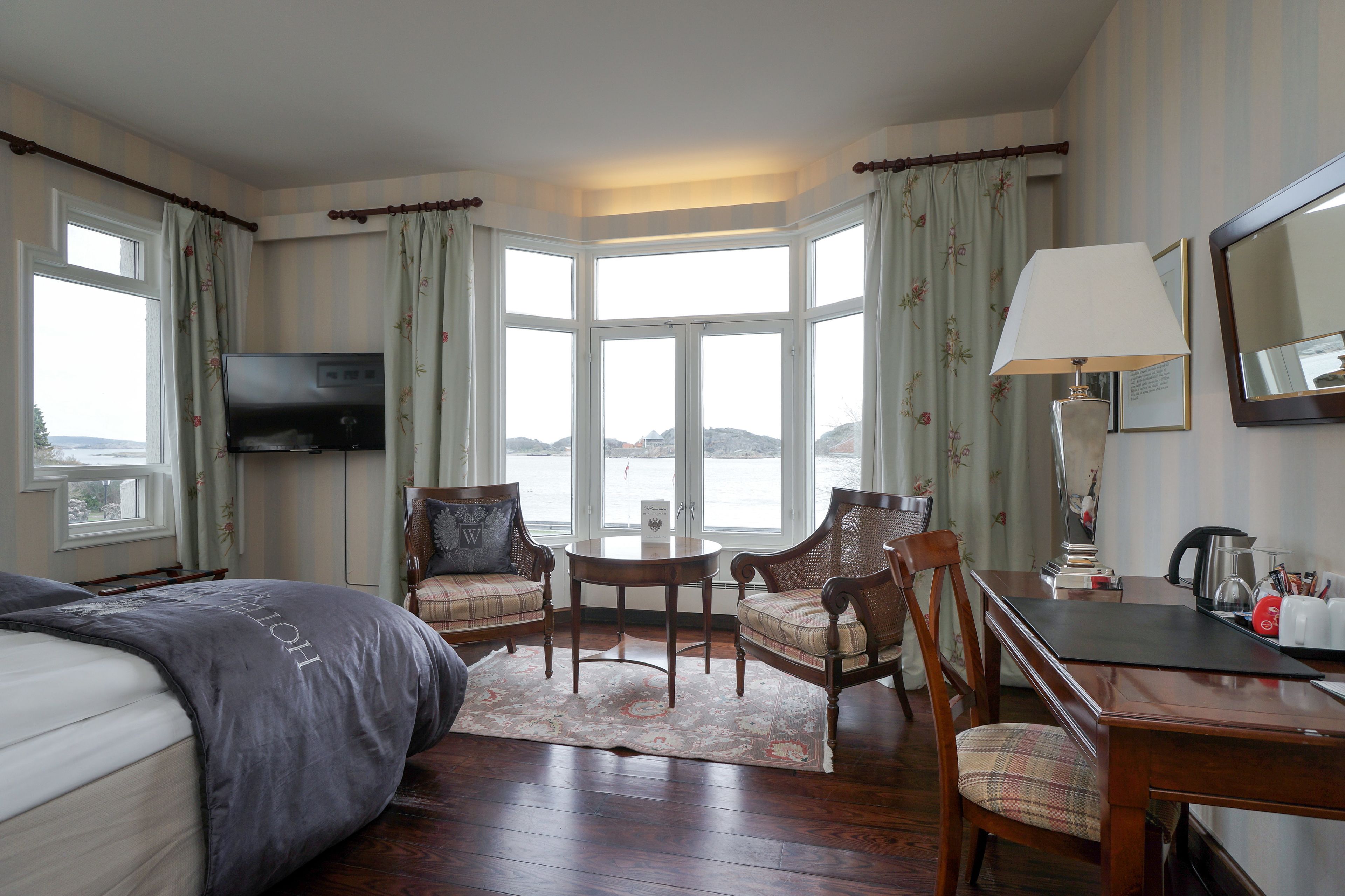 A hotel room with a bed, two armchairs, a small table, a desk, a lamp, a TV, and a large window with curtains.