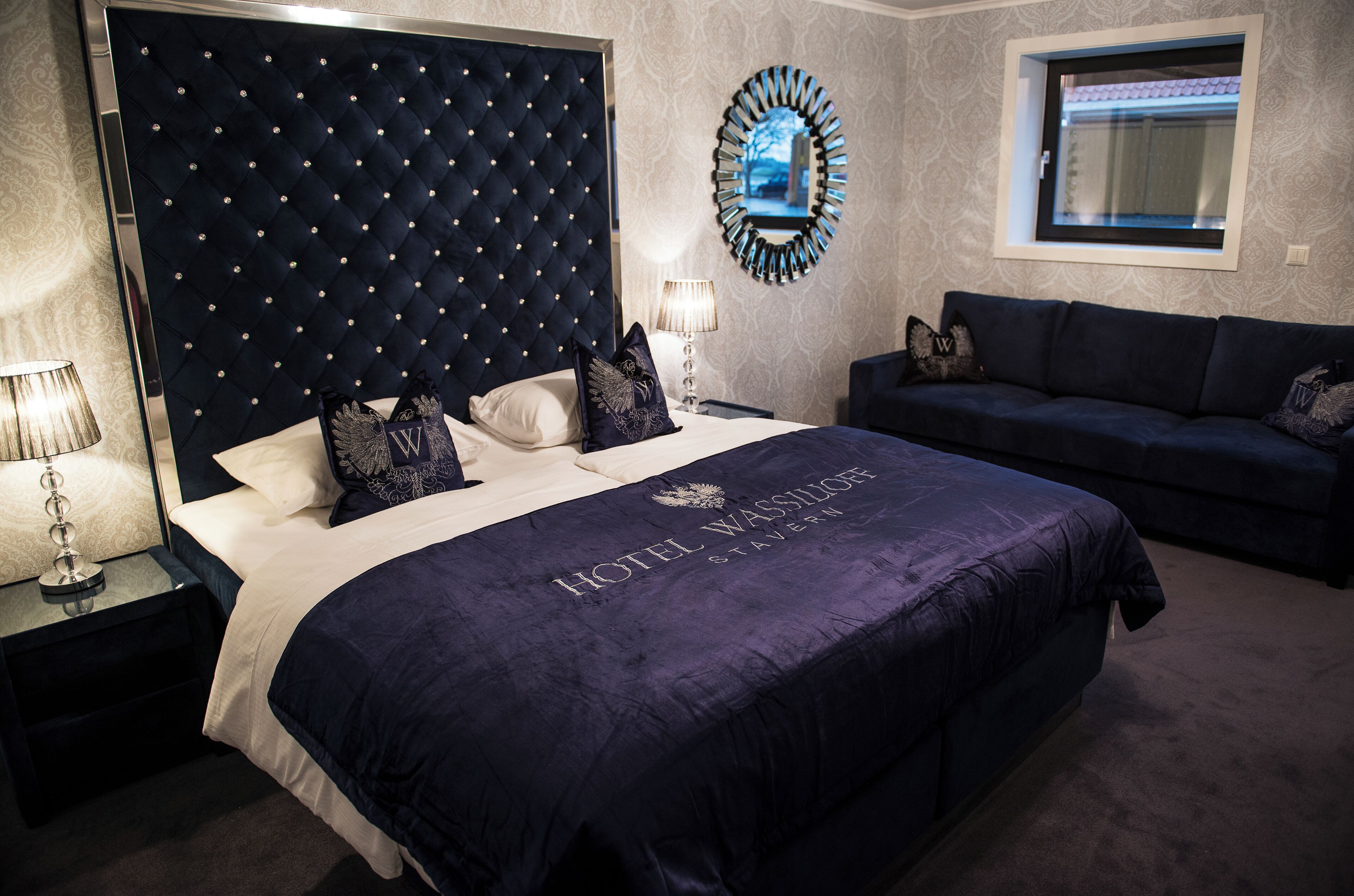 A hotel room with a large bed, a dark blue headboard, a sofa, a mirror, and a window.