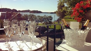 A table with champagne glasses and a bottle of Moët & Chandon champagne, set against a scenic view of a lake and red flowers. - Hotel Wassilioff - Unike Hoteller (Larvik)