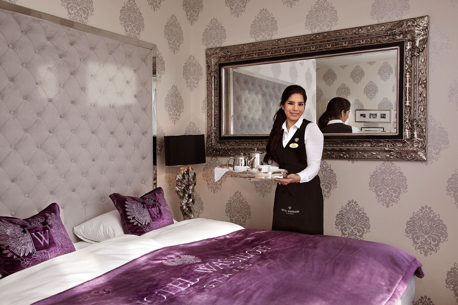 Una habitación de hotel con cabecera capitonada, un espejo grande y una cama con ropa de cama morada.