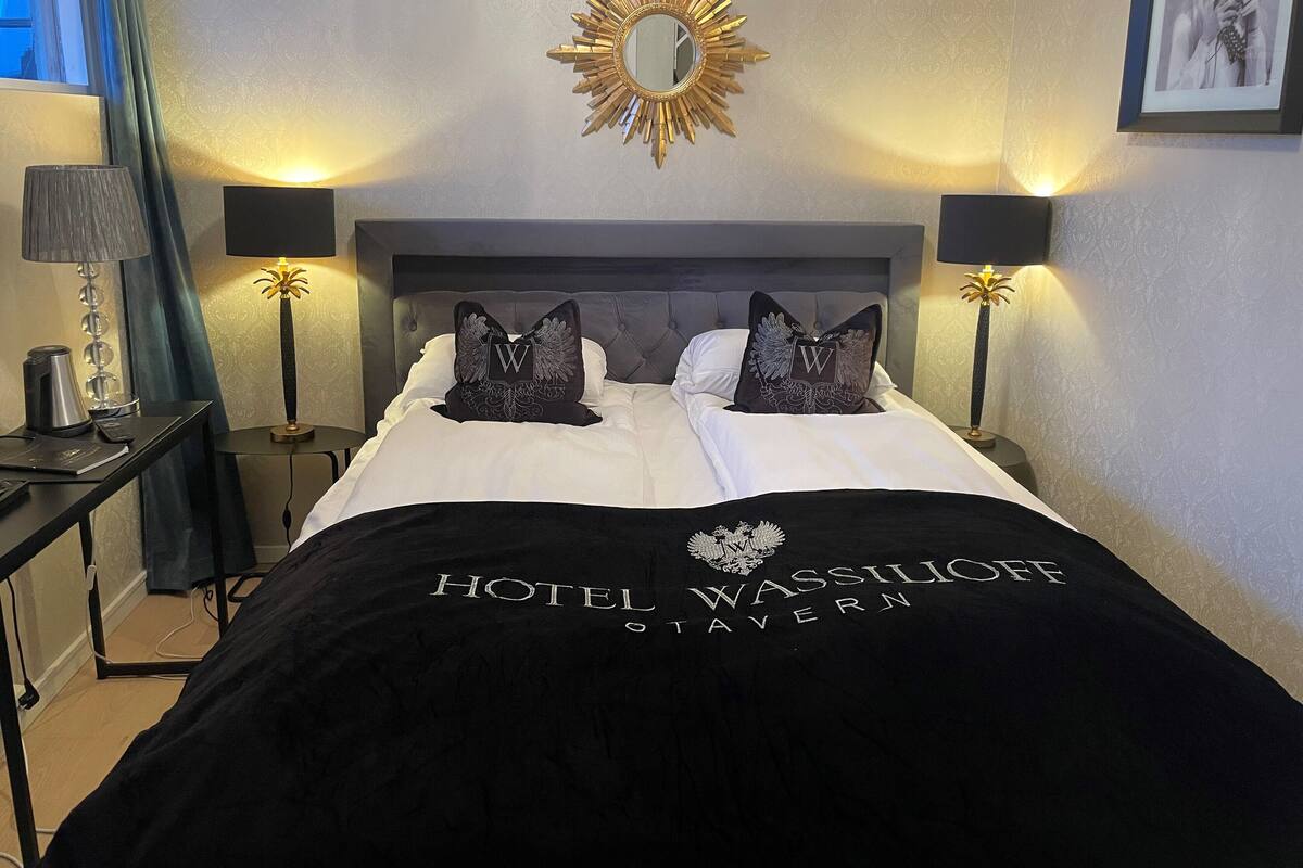 A hotel room with a bed, two pillows with a 'W' emblem, a gold mirror, and two lamps.
