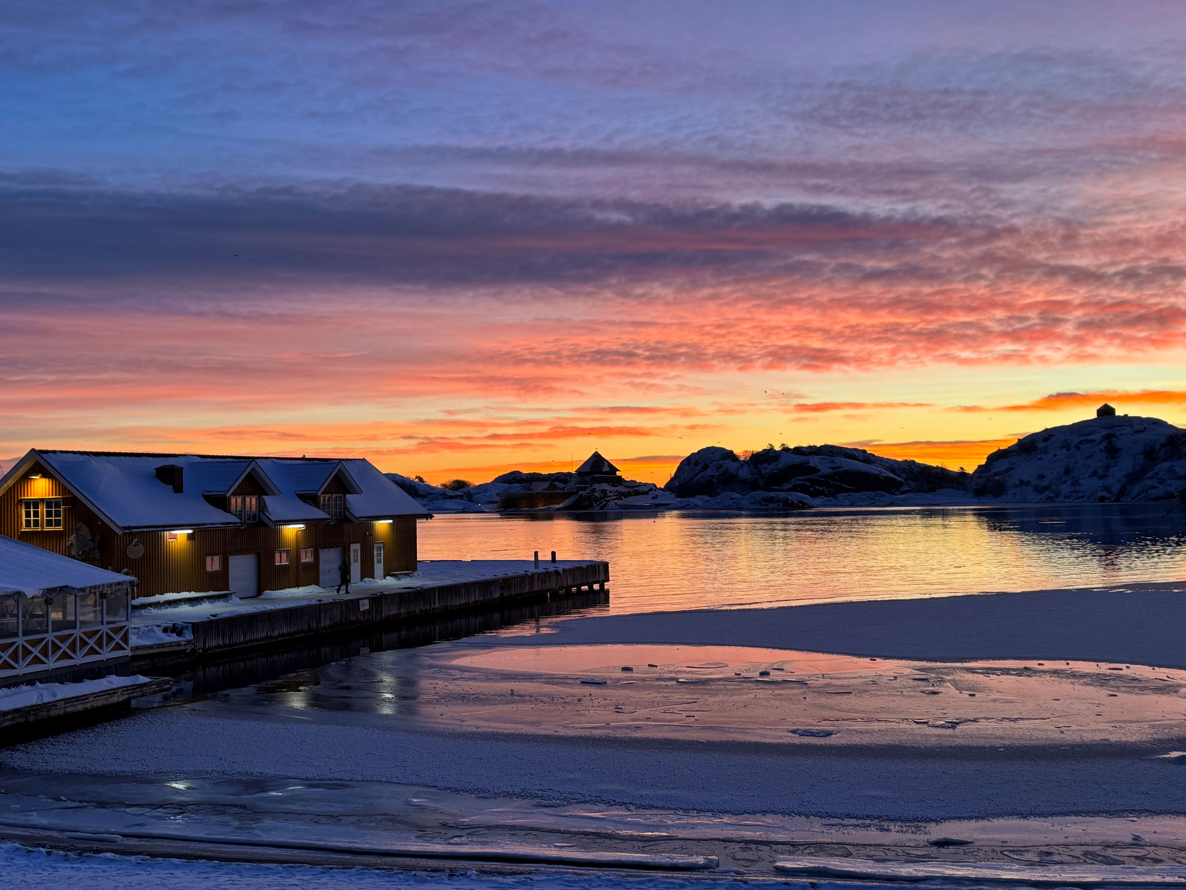 A waterfront property with a dock, snow-covered buildings, and a sunset.